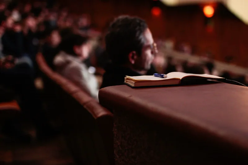 An open notebook rests on an auditorium seat with an audience blurred in the background.